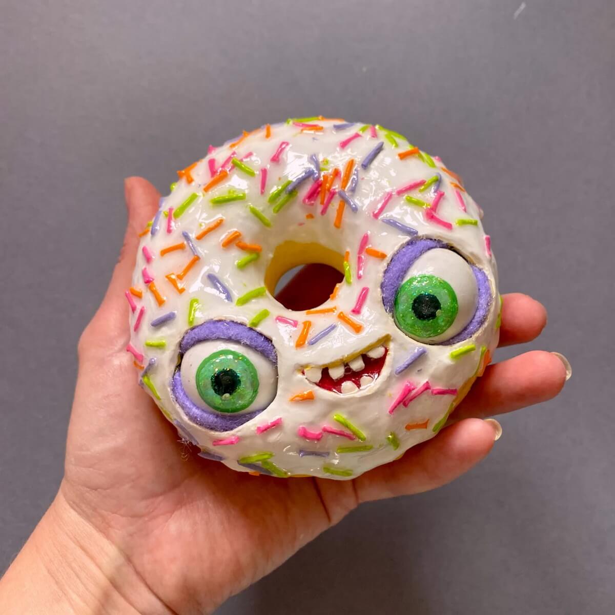 Papier-mâché donut with colorful sprinkles and funny face – handmade craft held in hand