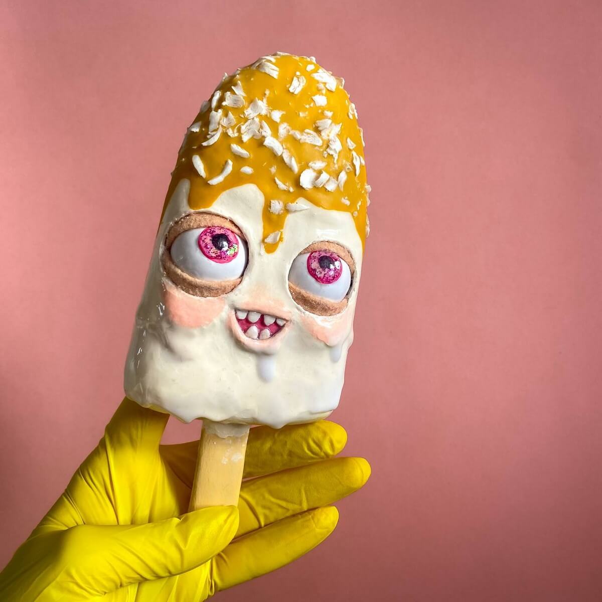 Yellow and white papier-mâché ice cream figure with expressive face – handmade craft held in yellow gloves on pink background