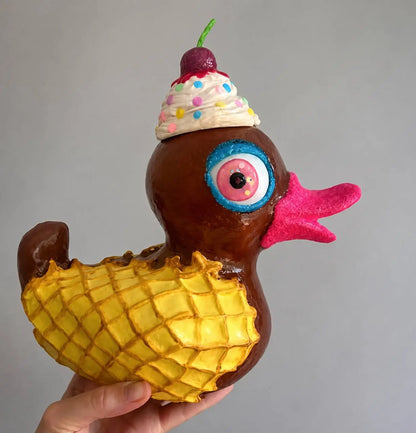A hand holds a whimsical rubber duck sculpture shaped like a chocolate duck with a yellow waffle shell body, a pink beak, a large blue-rimmed eye, and a whipped cream ice cream cone hat topped with a cherry, against a plain gray background.
