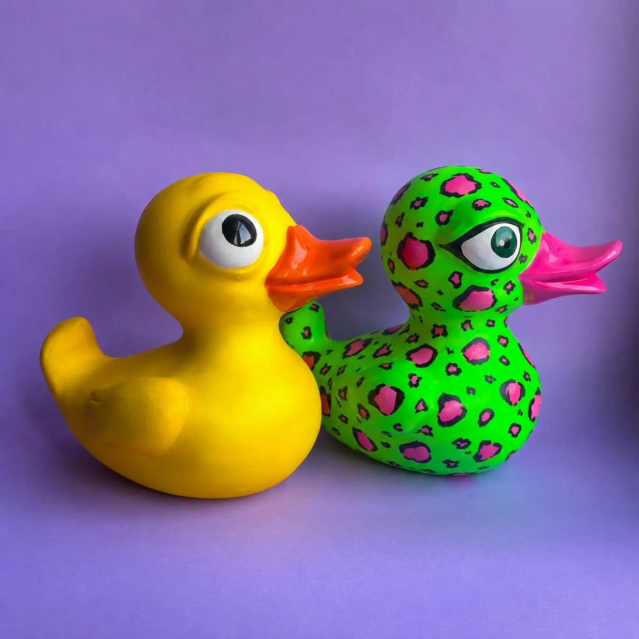 Two decorative rubber ducks on a solid purple background. The duck on the left is solid yellow with a bright orange beak and large cartoon eyes.