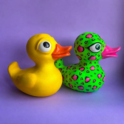 Two decorative rubber ducks on a solid purple background. The duck on the left is solid yellow with a bright orange beak and large cartoon eyes.