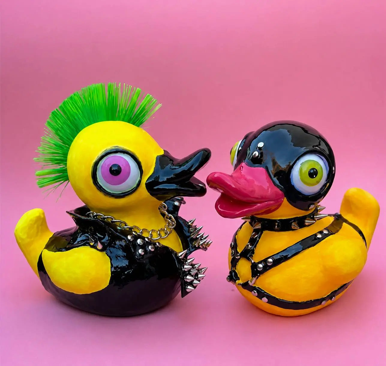 Two novelty rubber ducks styled in punk fashion, one with a green mohawk and black studded jacket, the other wearing a black mask and harness, set against a solid pink background.