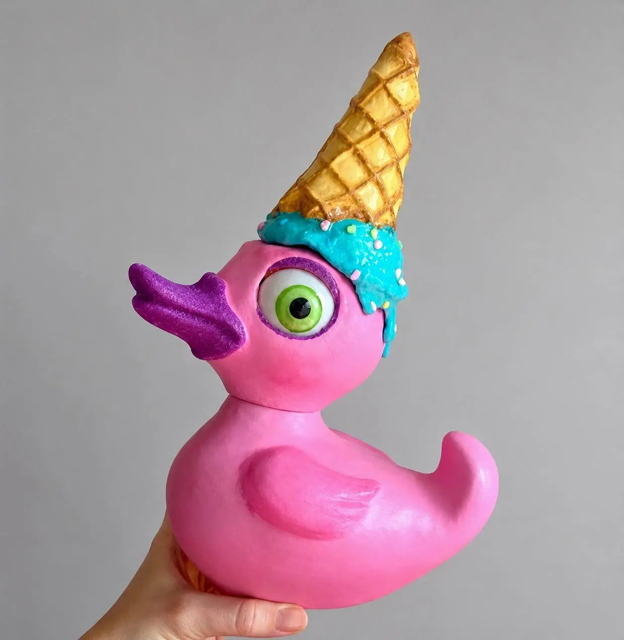 A pink rubber duck with a purple beak and a melting ice cream cone hat, held by a hand against a plain gray background.