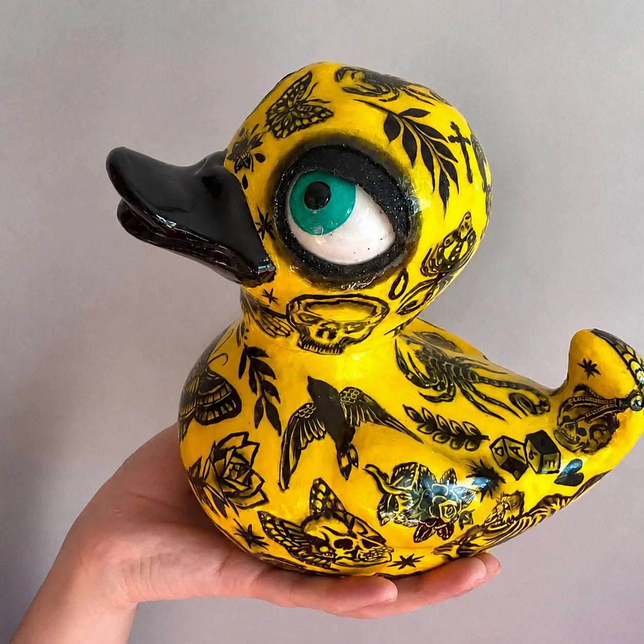 A hand holds a yellow rubber duck decorated with black tattoo-style illustrations, including skulls, butterflies, birds, and a large turquoise and white eye on the head, against a plain light gray background.
