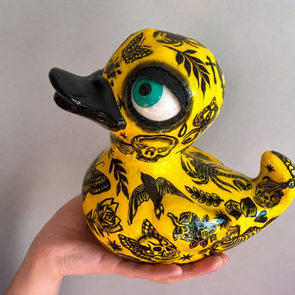A hand holds a yellow rubber duck decorated with black tattoo-style illustrations, including skulls, butterflies, birds, and a large turquoise and white eye on the head, against a plain light gray background.