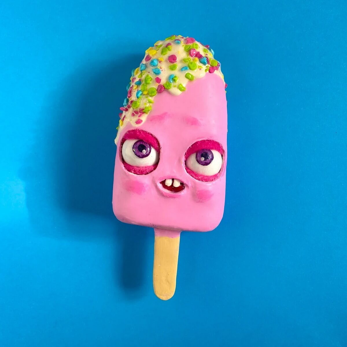Pink papier-mâché ice cream on a stick with funny face and candy topping – creative handmade sculpture on blue background