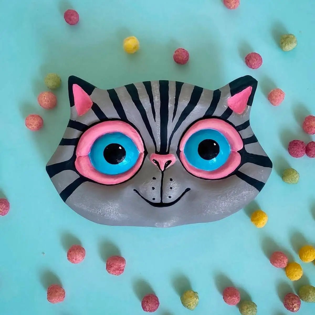 Gray cat wall sculpture surrounded by colorful pom-poms on blue surface